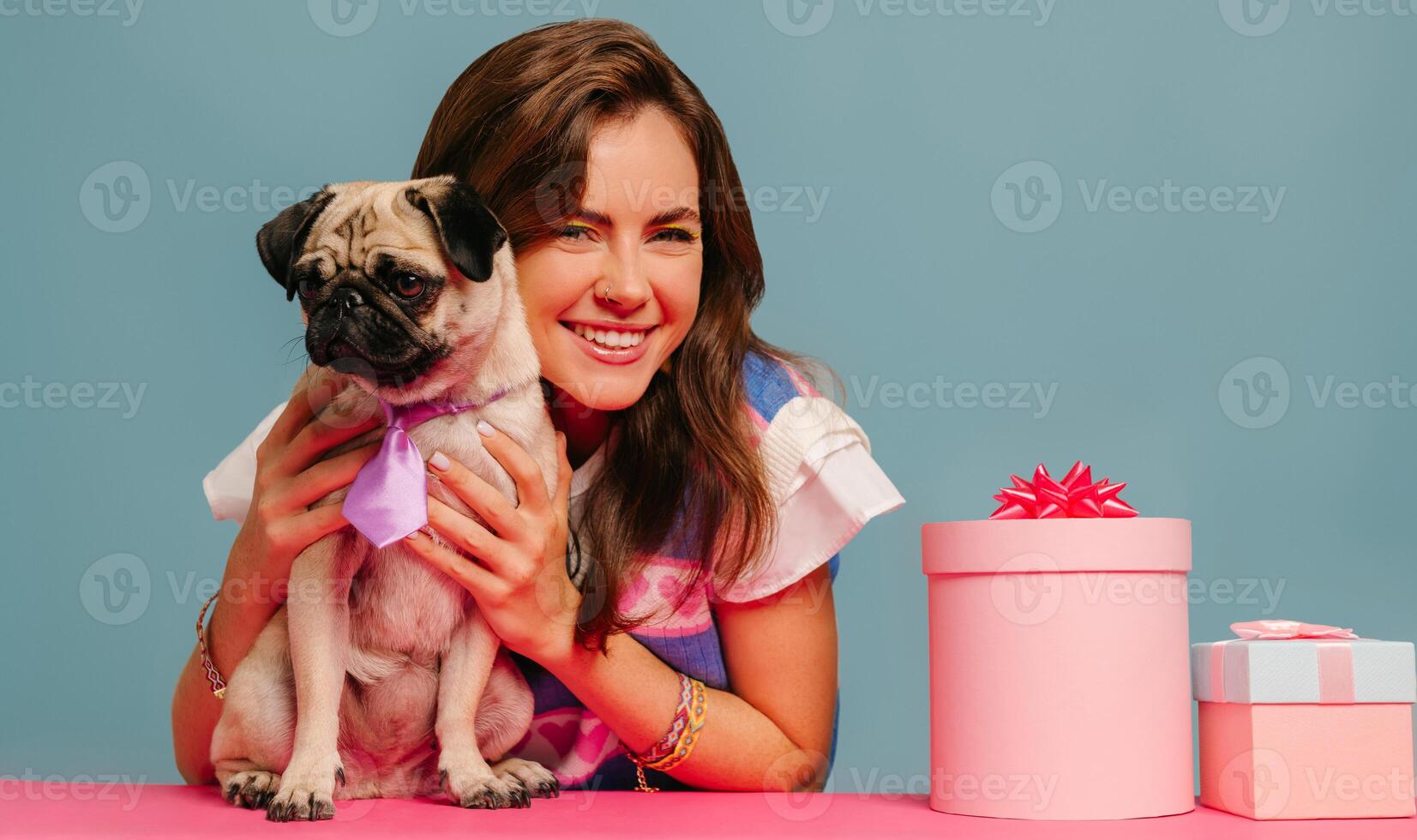 Attractive young woman petting cute pug dog and smiling while sitting at the pink desk on blue background photo