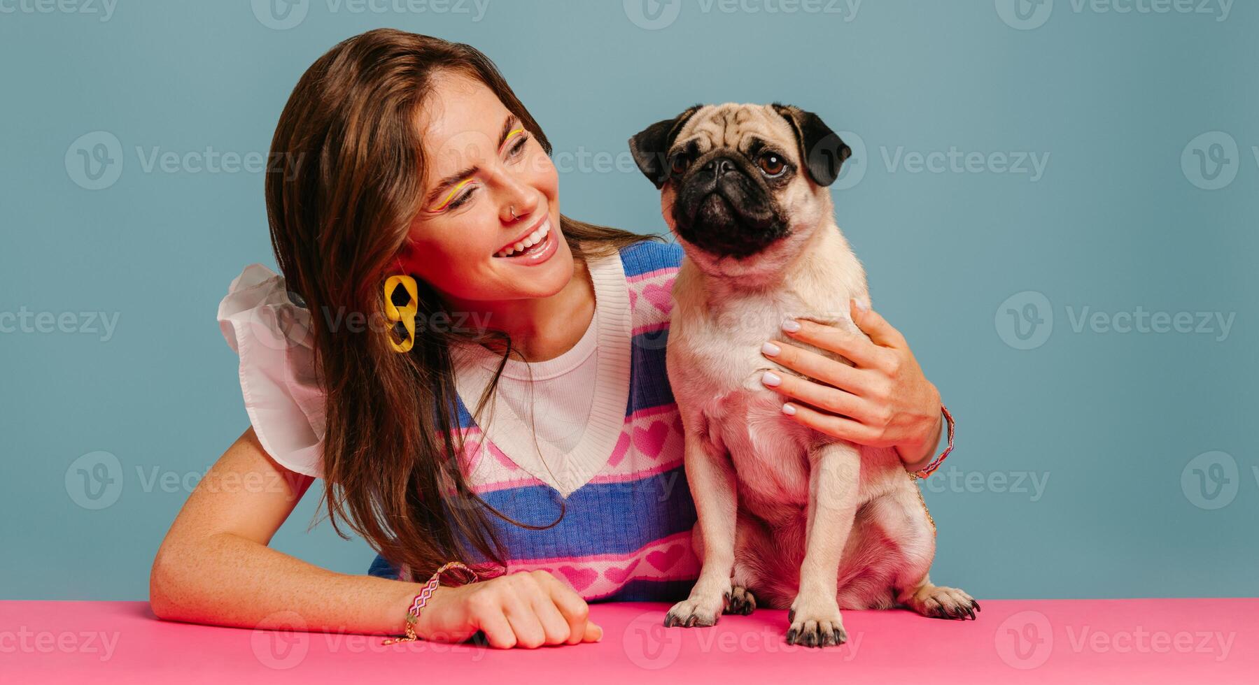 Attractive young woman hugging cute pug dog and smiling while sitting at the pink desk on blue background photo