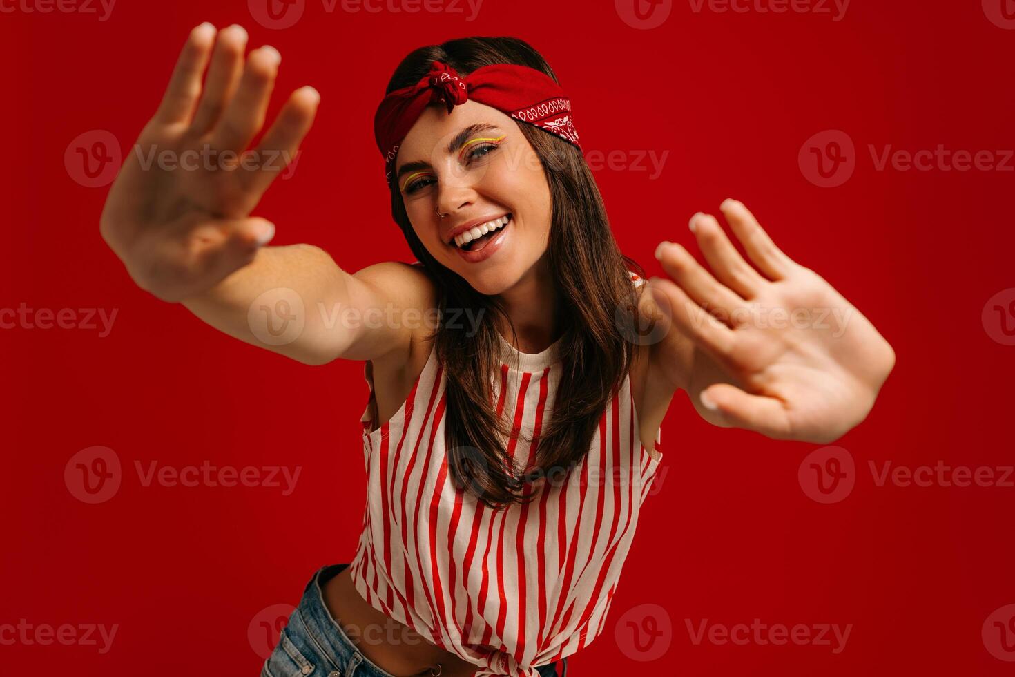 Joyful young hipster woman stretching out hands and smiling while standing on red background photo