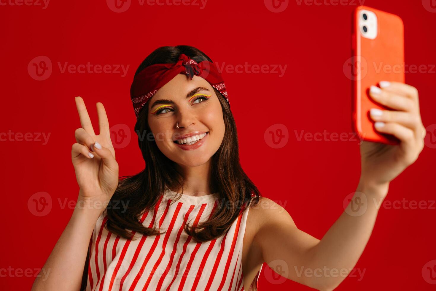 Joyful young hipster woman making selfie by mart phone and smiling while standing on red background photo