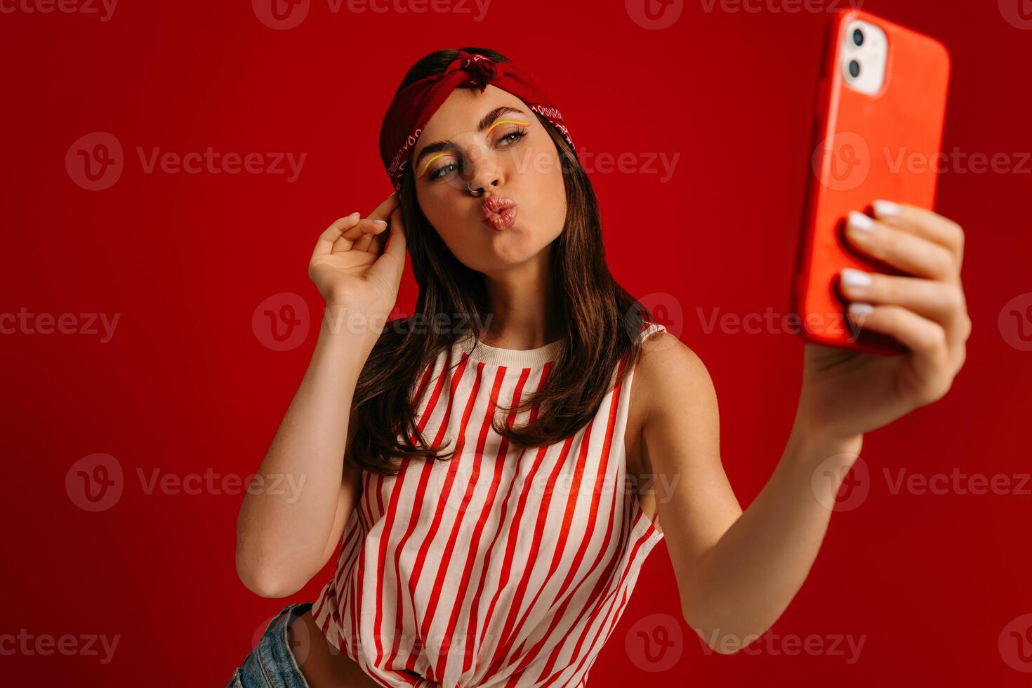 Playful young hipster woman making selfie by mart phone while standing on red background photo