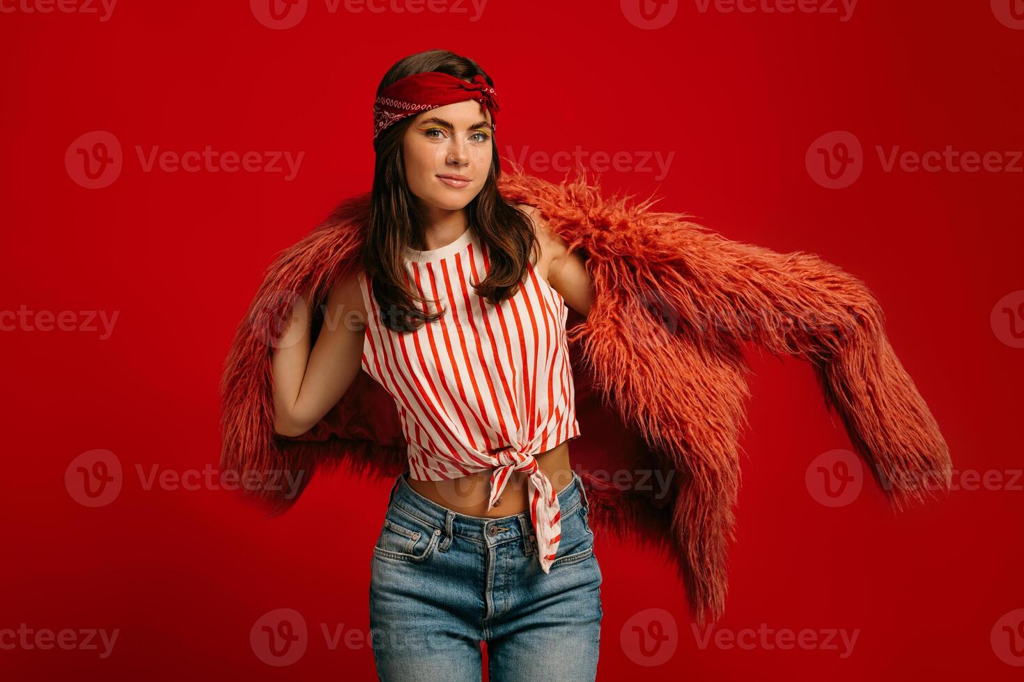 Fashionable young hipster woman wearing fluffy coat and smiling while standing on red background photo
