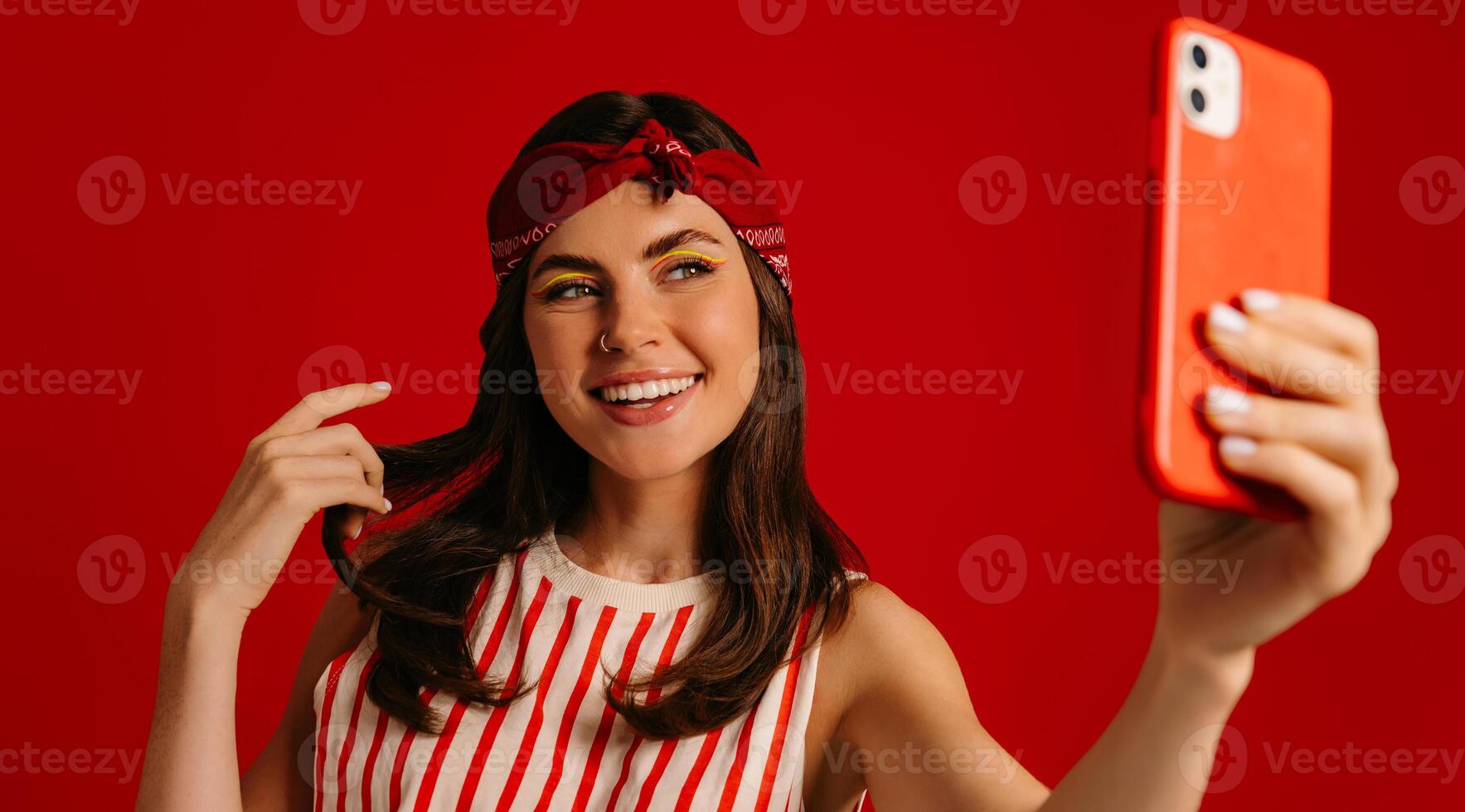 Beautiful young hipster woman making selfie by mart phone and smiling while standing on red background photo