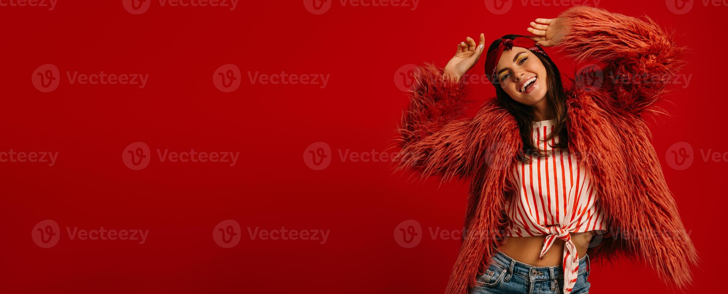 Fashionable young hipster woman wearing fluffy coat and dancing on red background photo