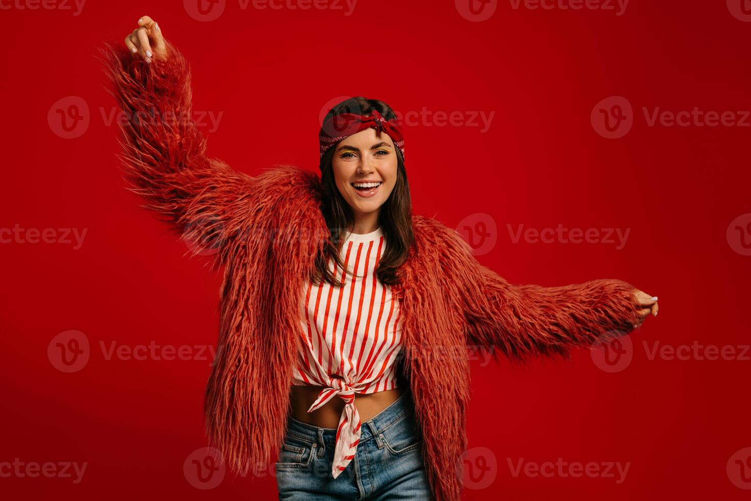 Fashionable young hipster woman wearing fluffy coat and dancing on red background photo