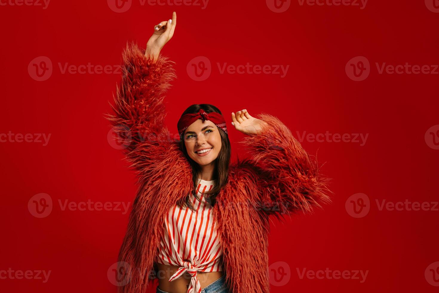 Joyful young hipster woman in fluffy coat dancing against red background photo