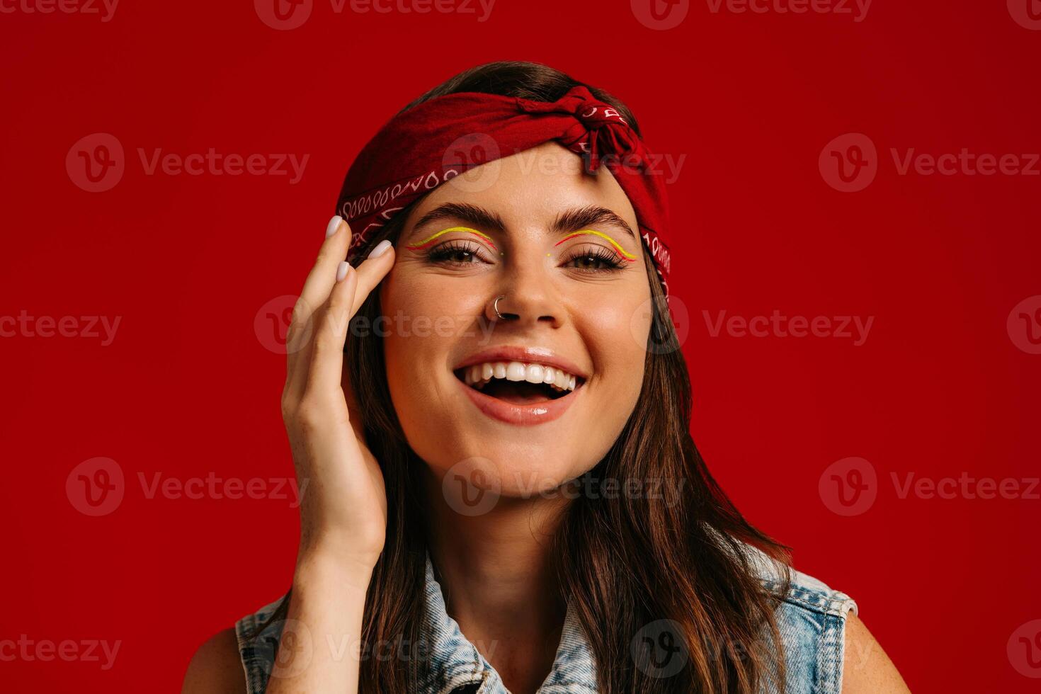 Cheerful young hipster woman with colorful make-up touching face and smiling on red background photo