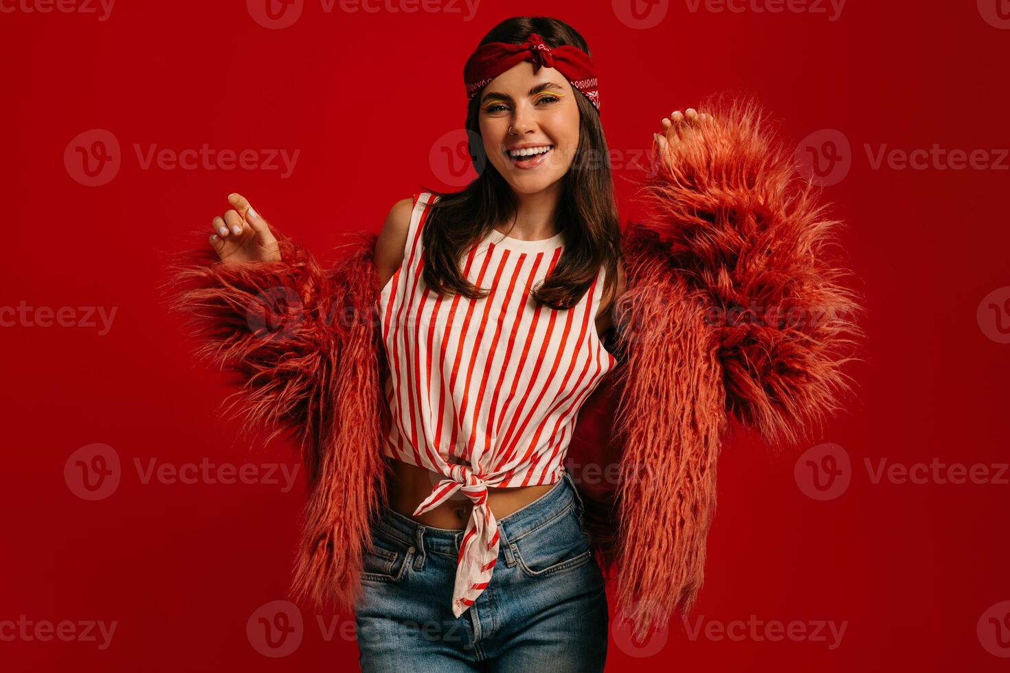 Joyful young hipster woman wearing fluffy coat dancing against red background photo