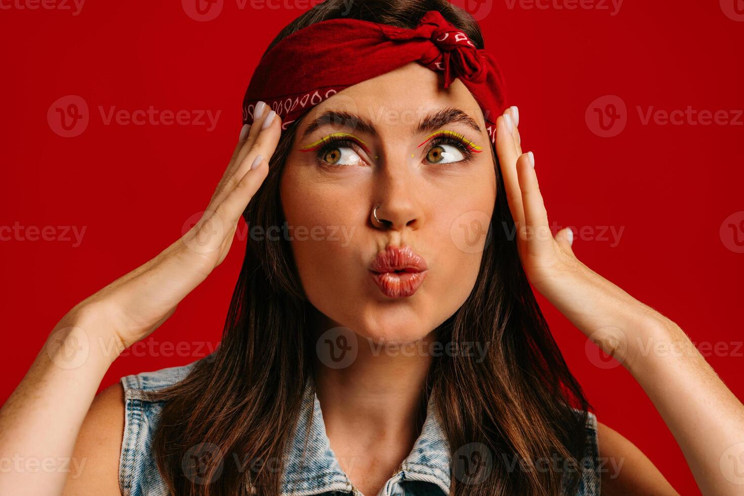 Beautiful young hipster woman with colorful make-up touching face and grimacing on red background photo