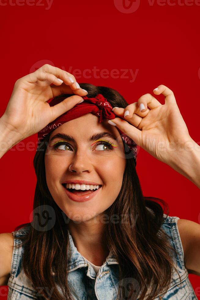 Attractive young hipster woman with colorful make-up adjusting bandana and smiling on red background photo