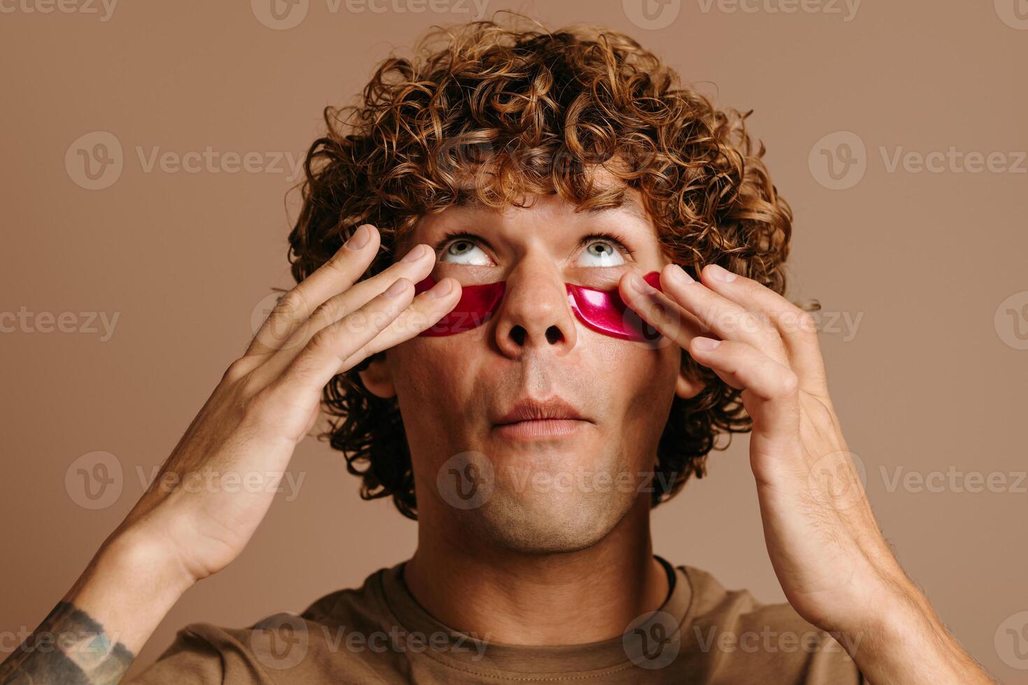 Handsome young curly man taking care of his skin while applying facial cosmetic mask on brown background photo