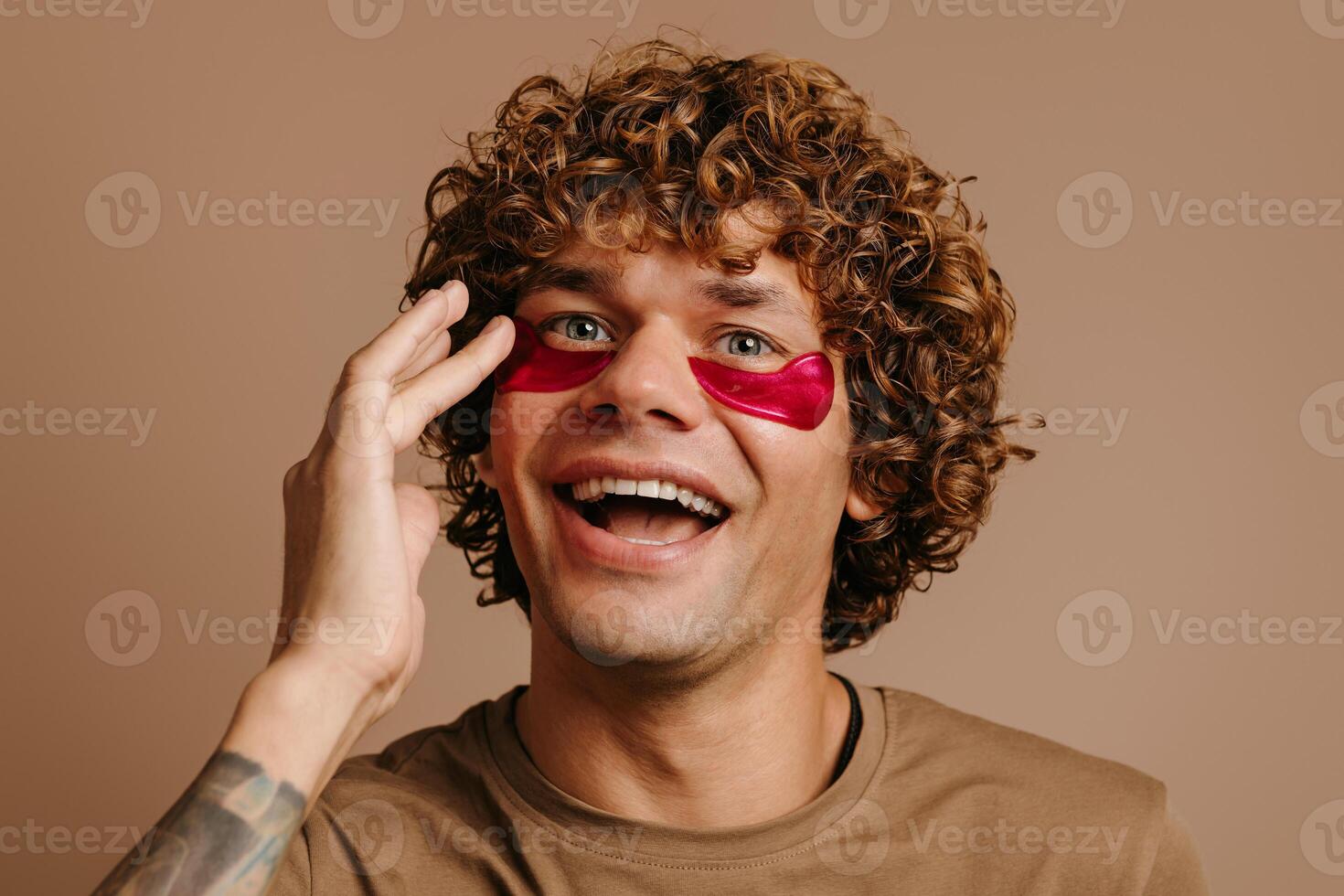 Happy young man taking care of his skin while applying anti-aging facial cosmetic mask on brown background photo
