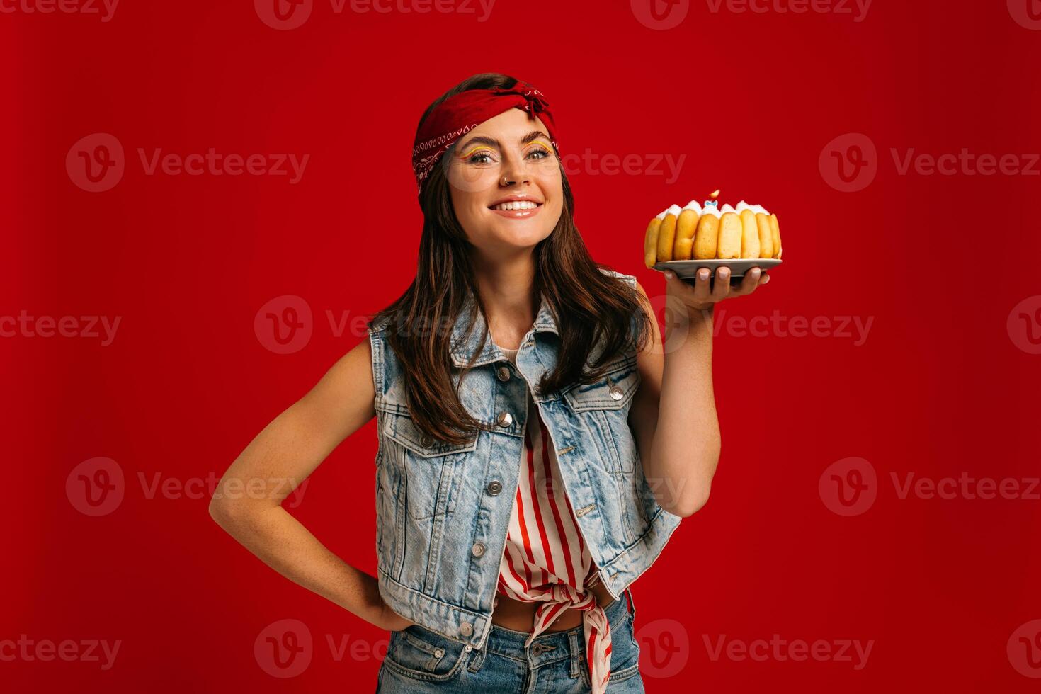 Happy young woman holding birthday cake and looking at camera with smile on red background photo