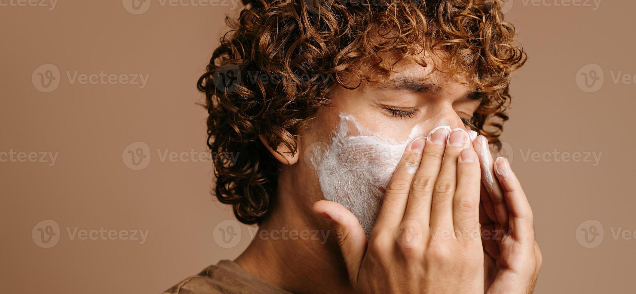 Handsome man enjoying beauty treatment while applying cream on face and standing on brown background photo