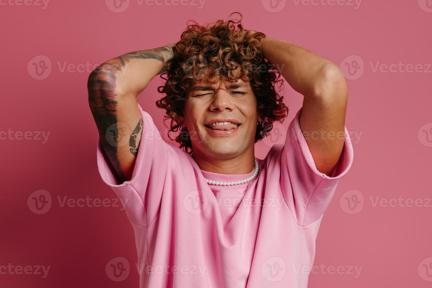 Playful young curly hair man holding head in hands and keeping eyes closed against pink background photo