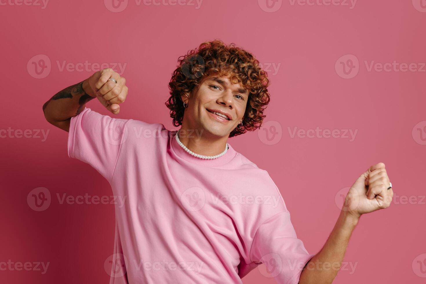 Happy young curly hair man dancing and smiling at camera against pink background photo