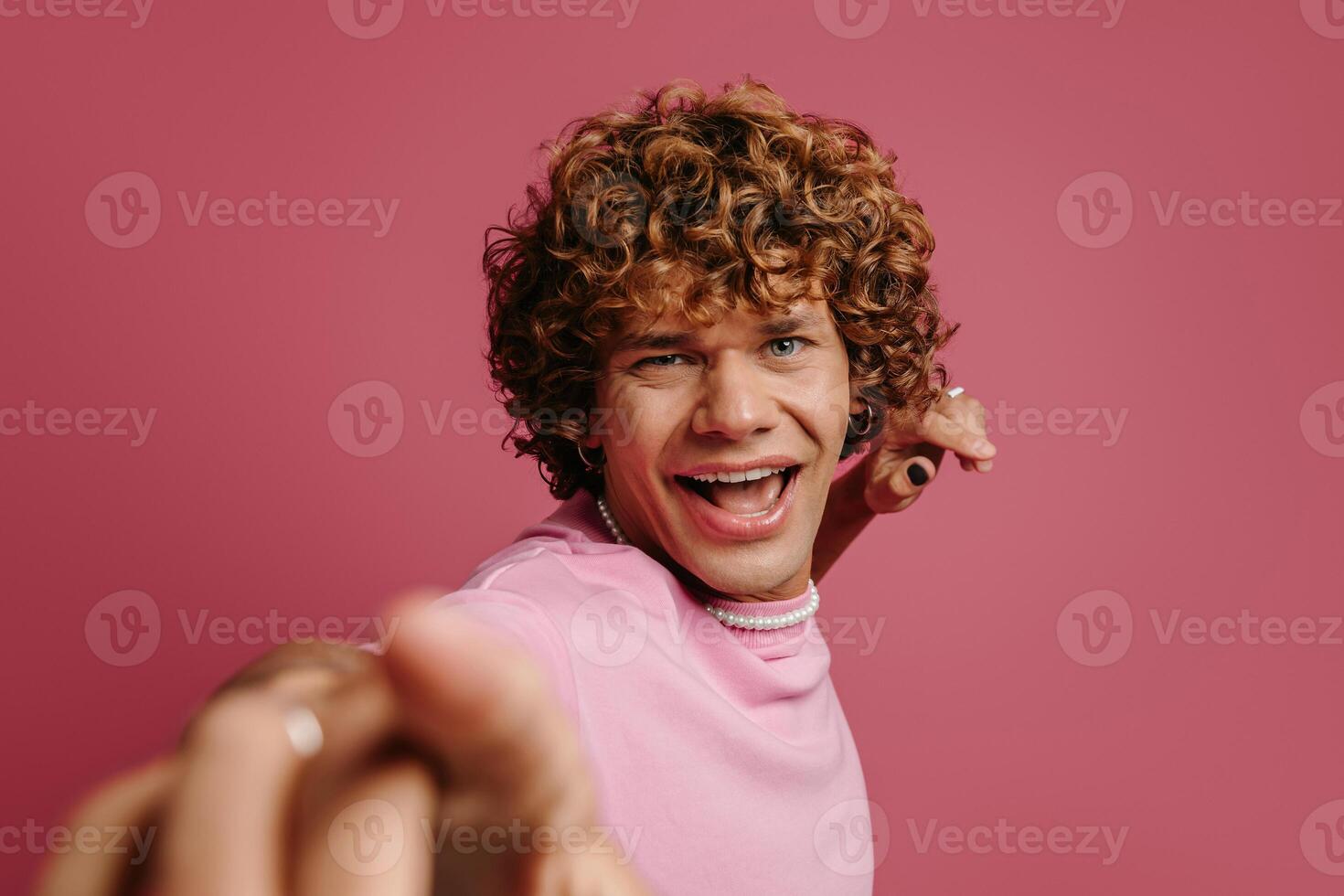 Playful young curly hair man gesturing and smiling at camera against pink background photo