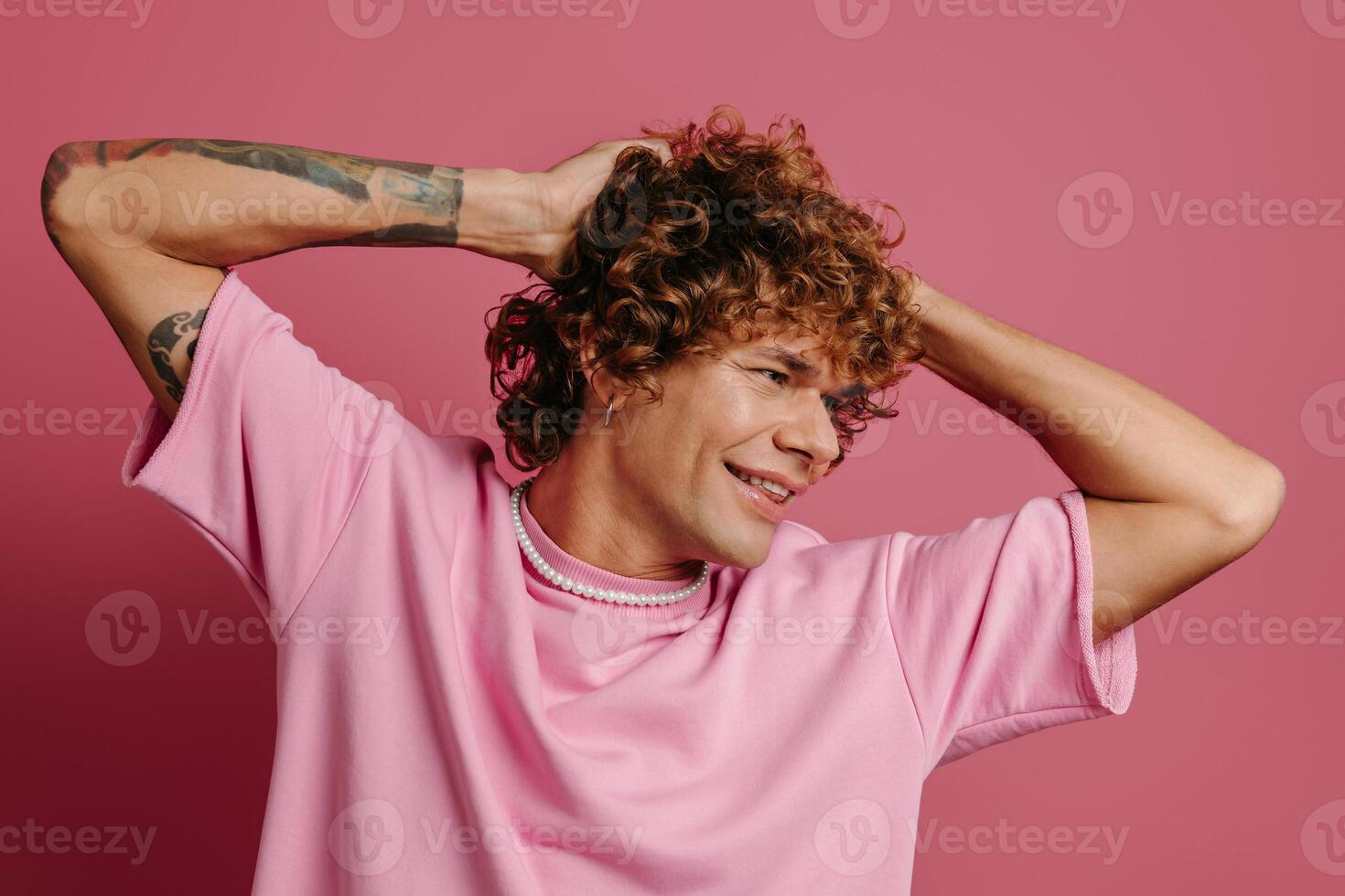 Fashionable young man holding hands in curly hair and smiling against pink background photo
