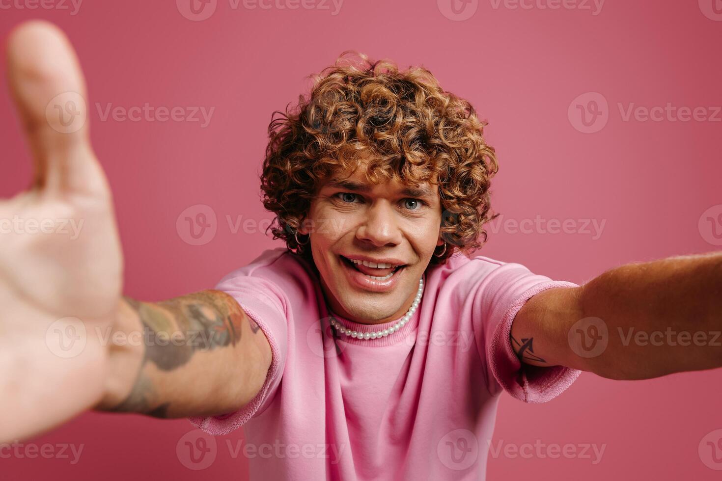 Handsome young curly hair man stretching out hands and smiling while making selfie against pink background photo