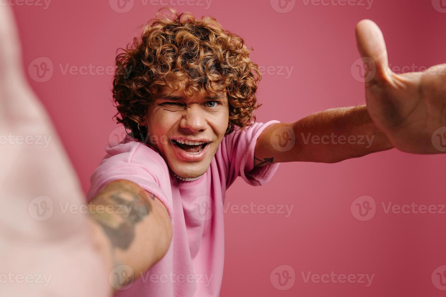 Playful young curly hair man stretching out hands and smiling while making selfie against pink background photo