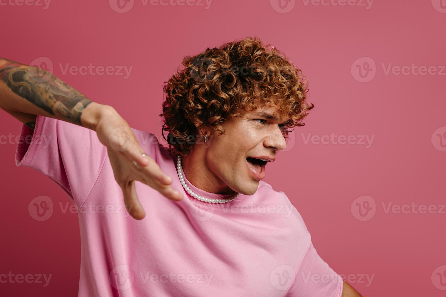 Happy young curly hair man gesturing and smiling at camera against pink background photo