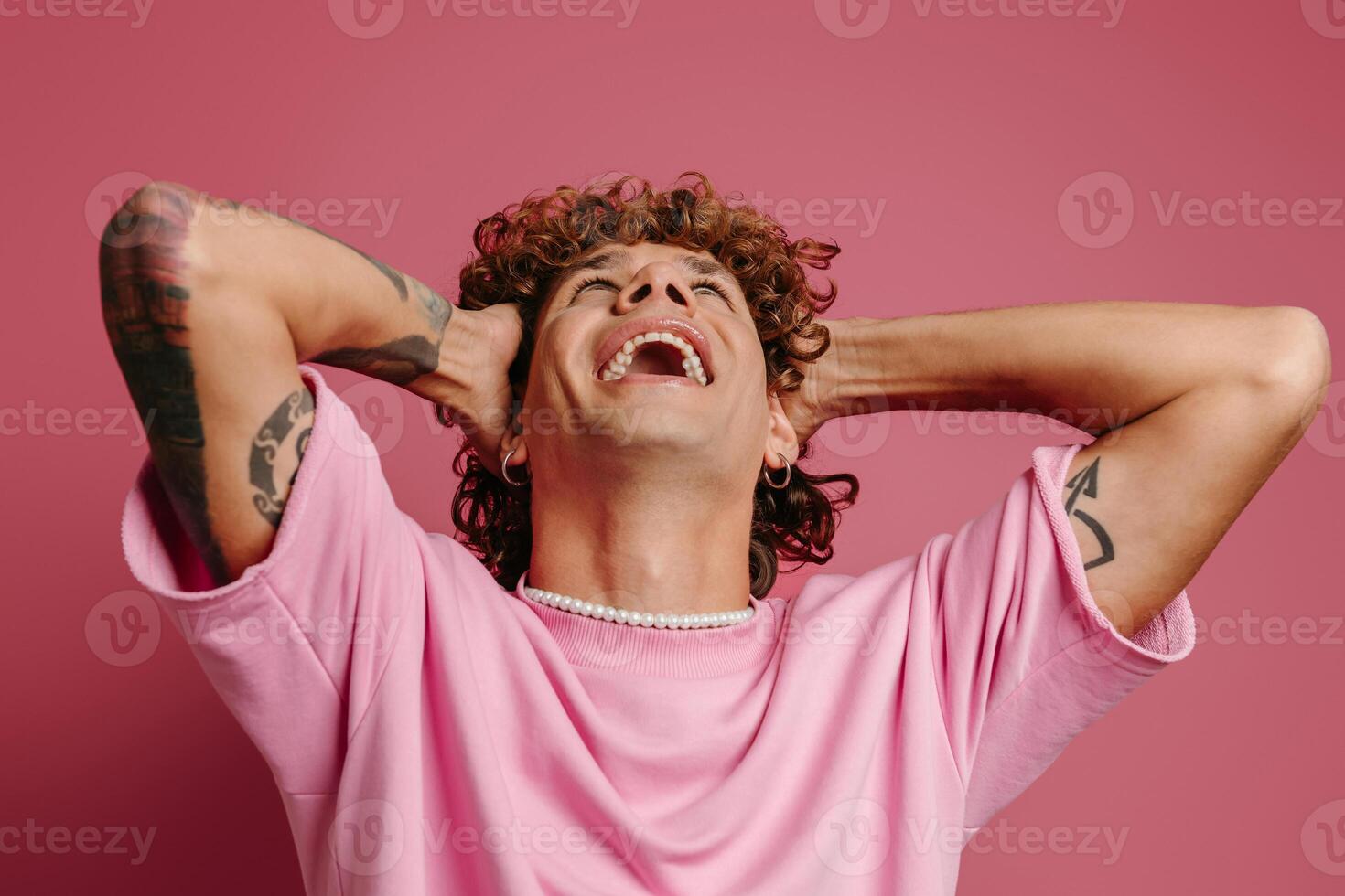 Excited young man holding hands in curly hair and smiling against pink background photo