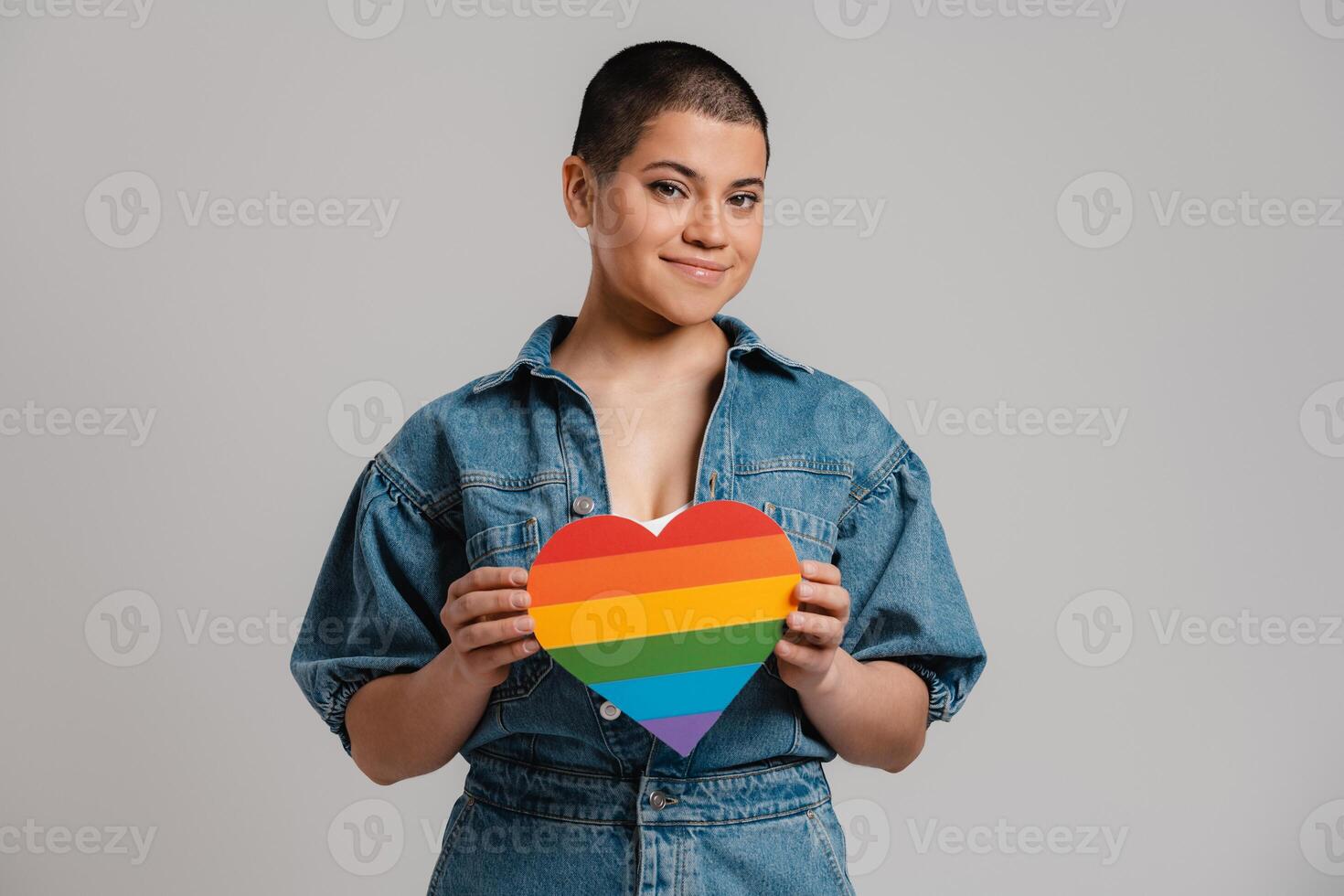 Beautiful young short hair woman holding heart shaped rainbow flag against grey background photo