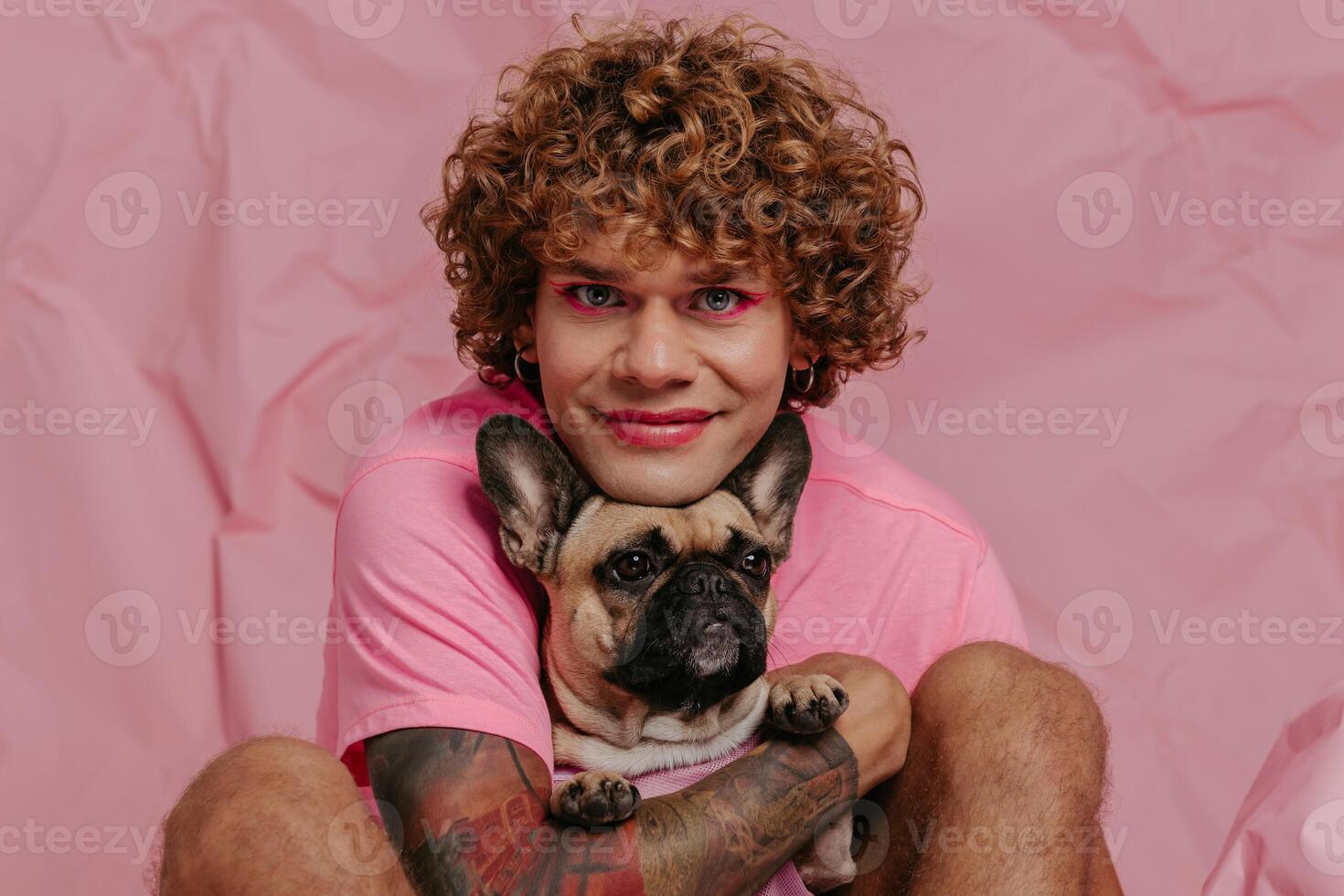 Handsome young gay man holding his cute dog and smiling while sitting against crumpled pink background photo