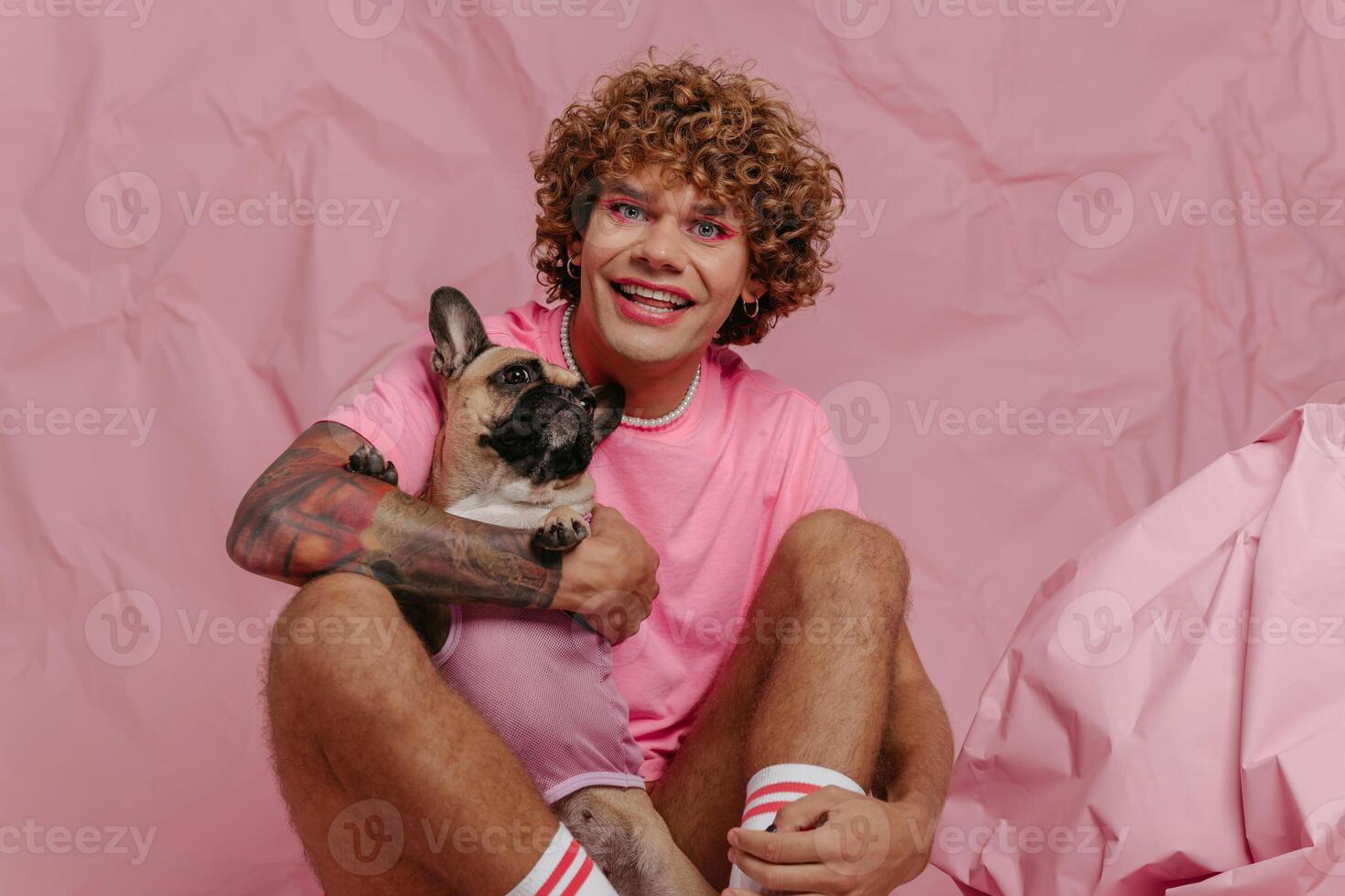 Stylish young gay man holding his cute dog and smiling while sitting against crumpled pink background photo