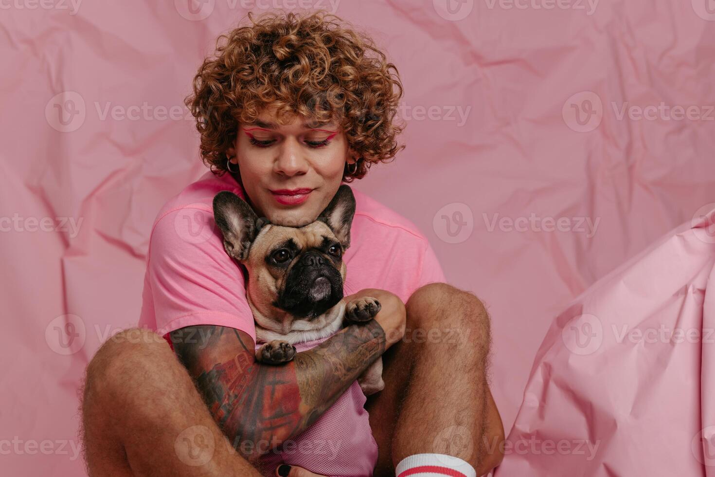 Stylish young gay man embracing his cute dog and smiling while sitting against crumpled pink background photo