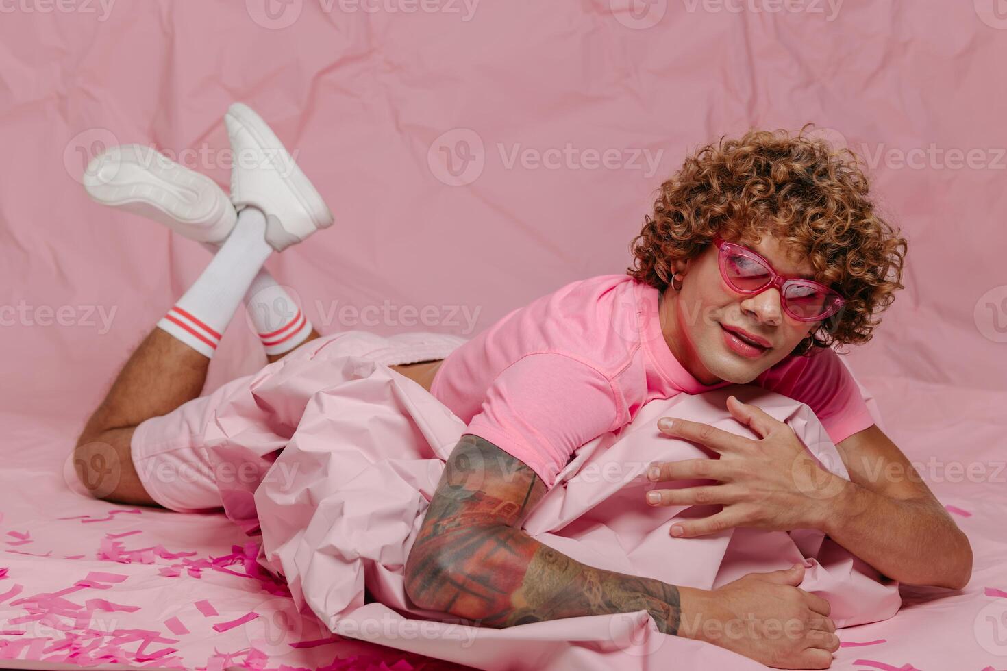 Stylish young gay man in trendy wear leaning on wad of crumpled pink paper while lying against colored background photo