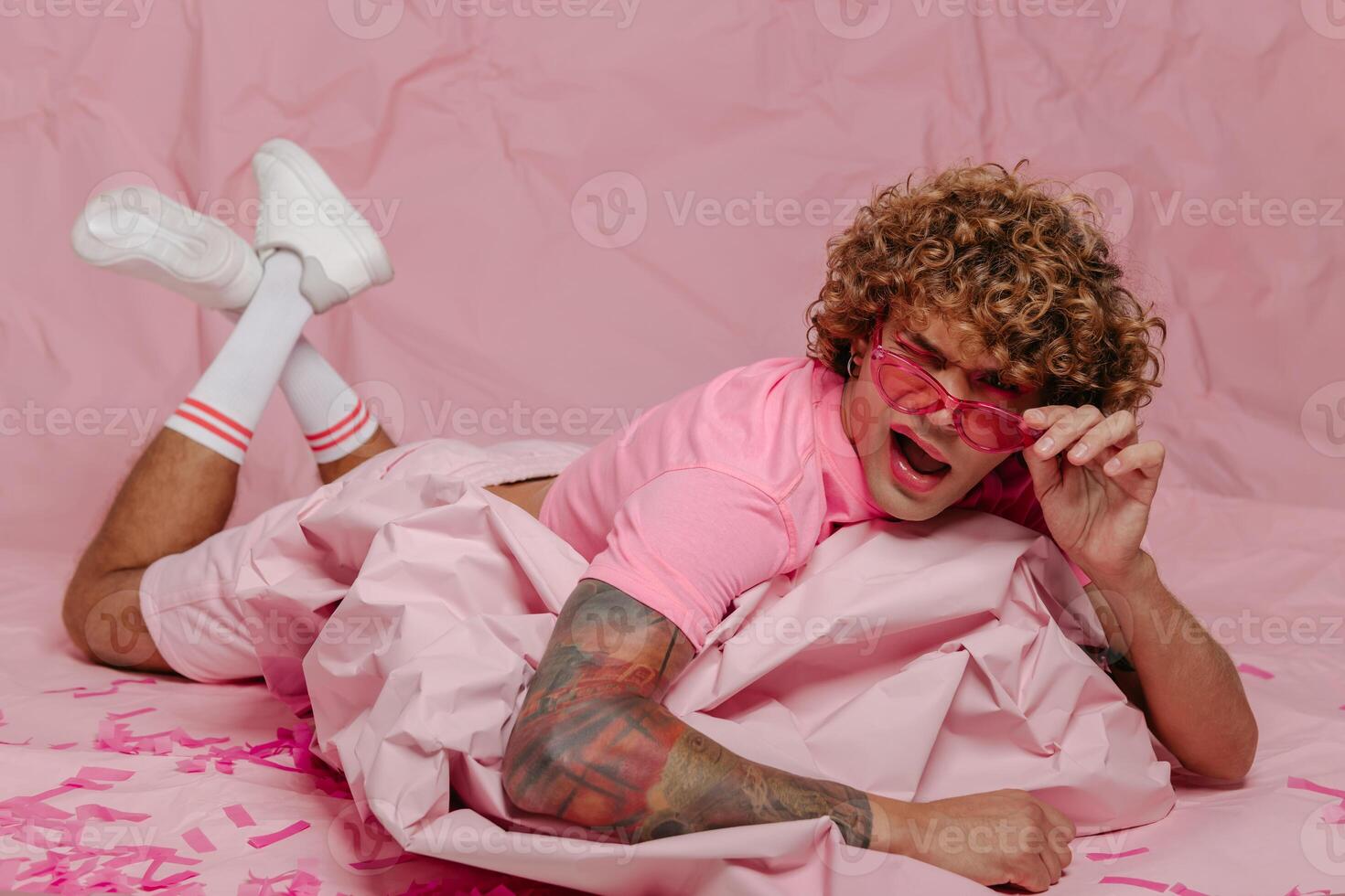 Stylish young gay man leaning on wad of crumpled pink paper while lying against colored background photo