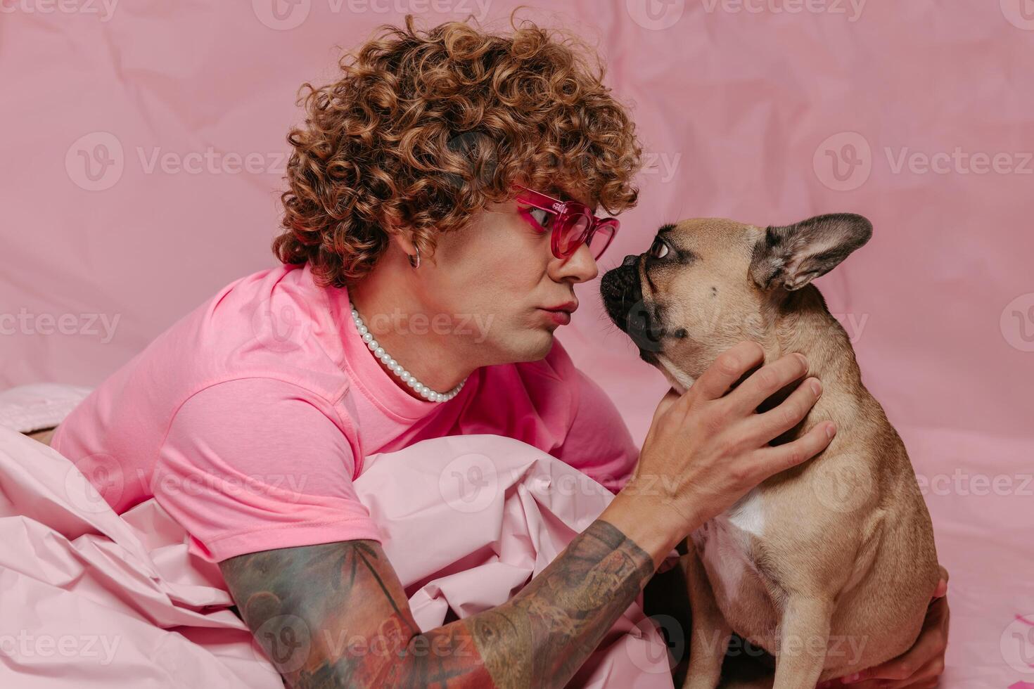 Stylish gay man leaning on wad of crumpled pink paper and petting dog while lying against colored background photo