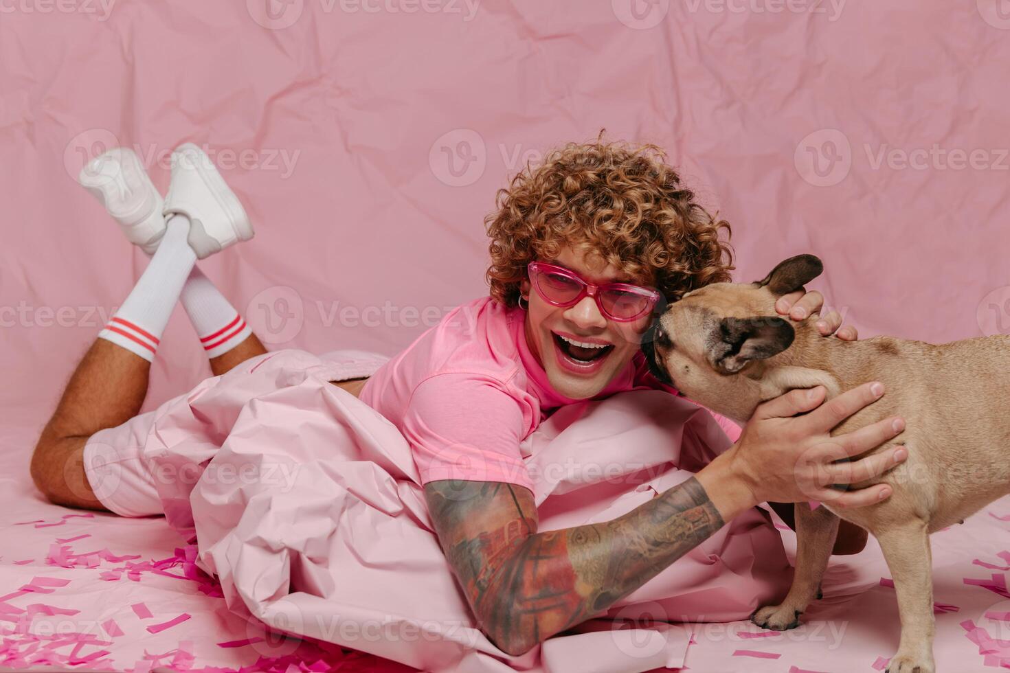 Stylish gay man leaning on wad of crumpled pink paper and petting dog while lying against colored background photo