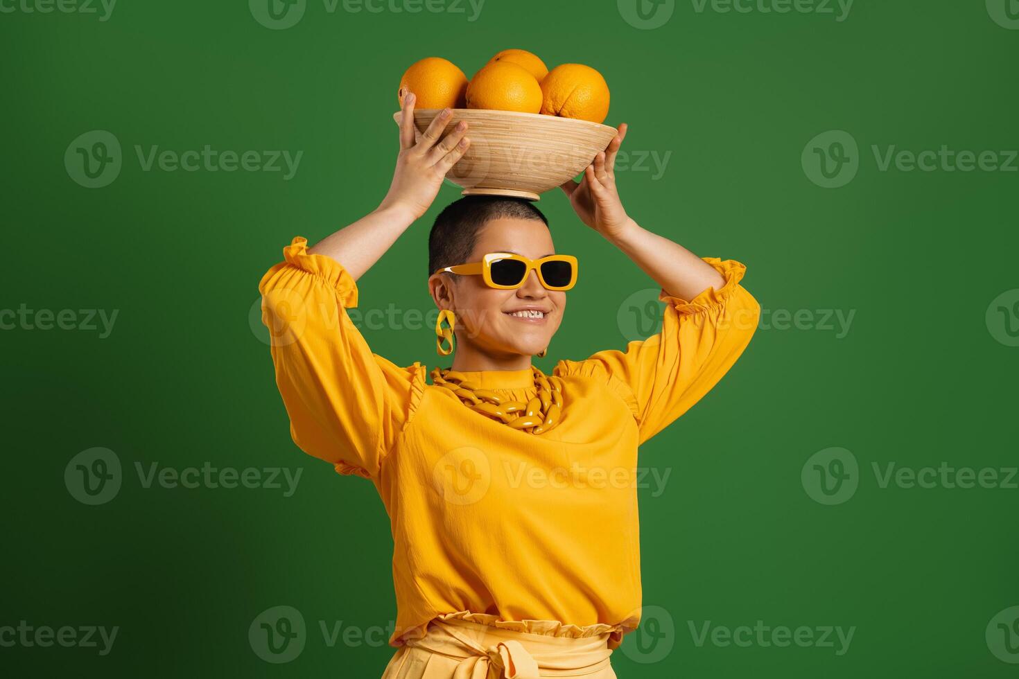 Happy young short hair woman carrying plate with oranges on head against green background photo