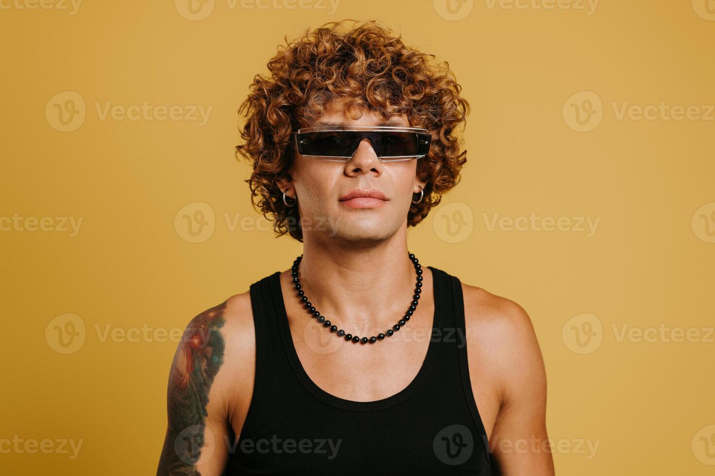 Fashionable curly young man in trendy glasses carrying fluffy coat on shoulder against yellow background photo