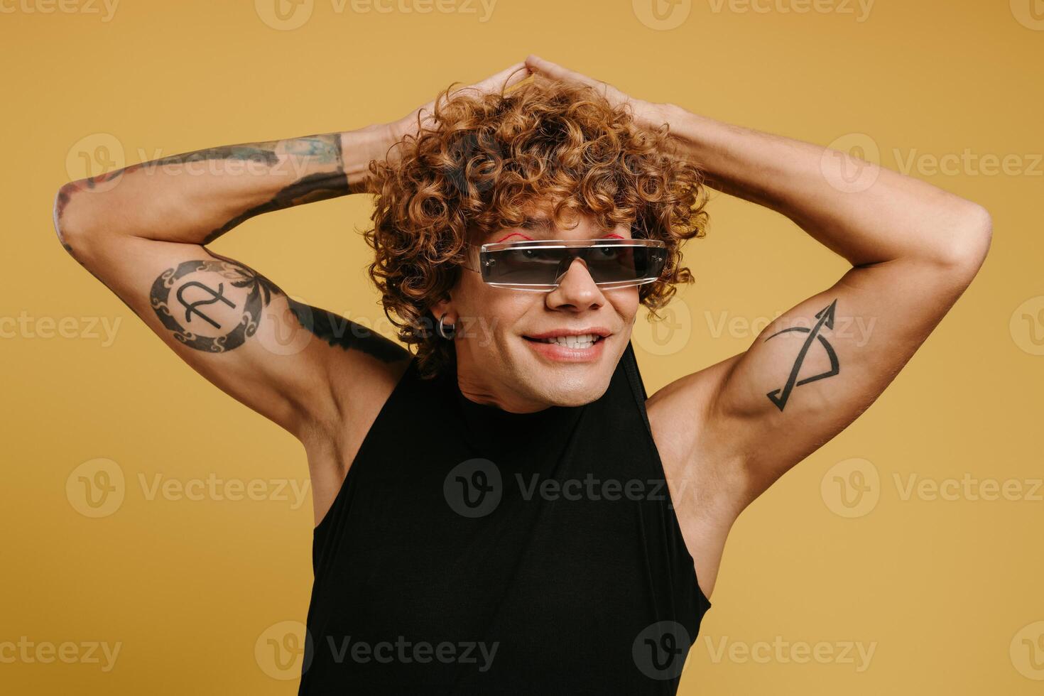 Joyful curly young man in trendy glasses playing with his tank top against yellow background photo