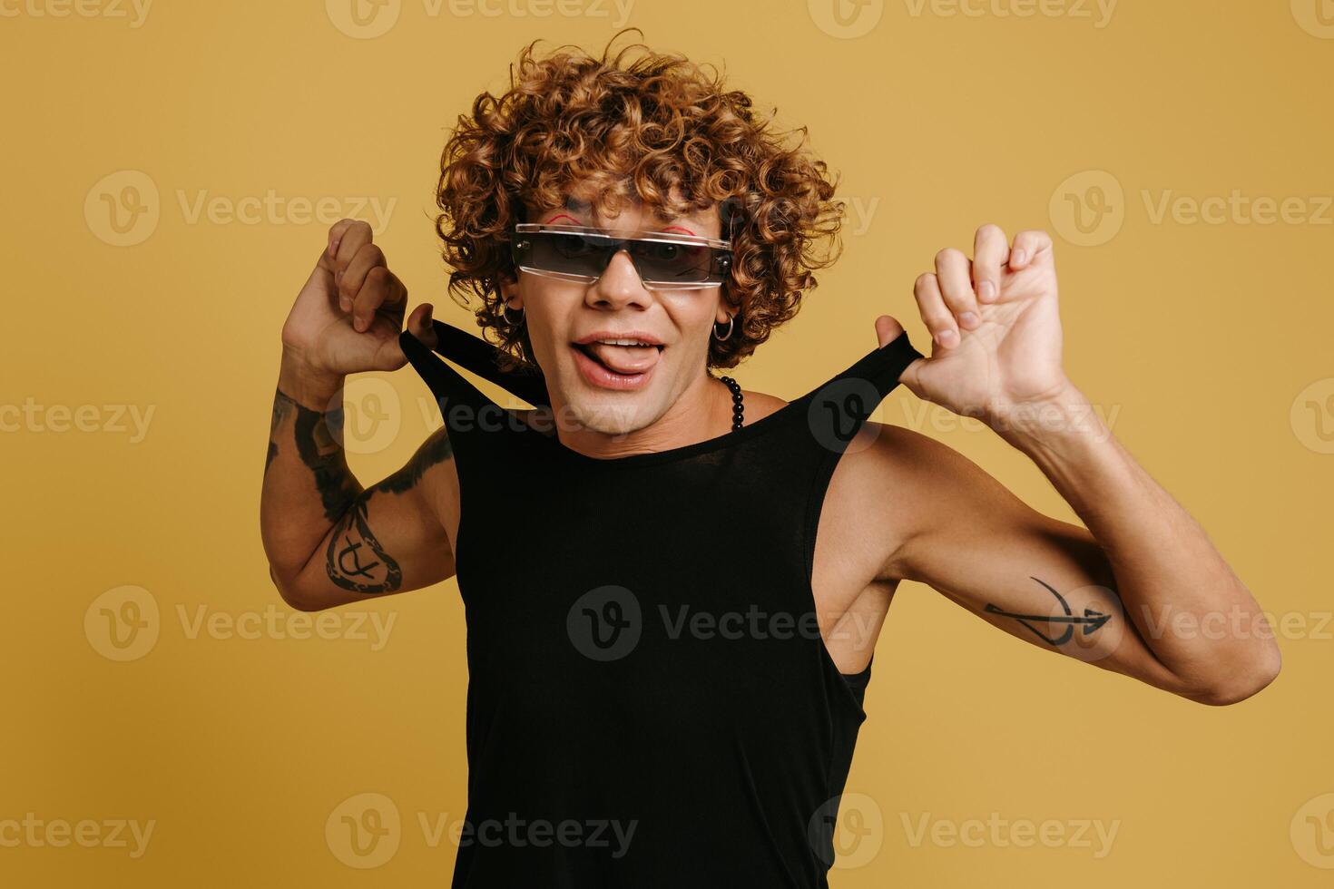 Playful curly young man in trendy glasses stretching tank top and smiling against yellow background photo