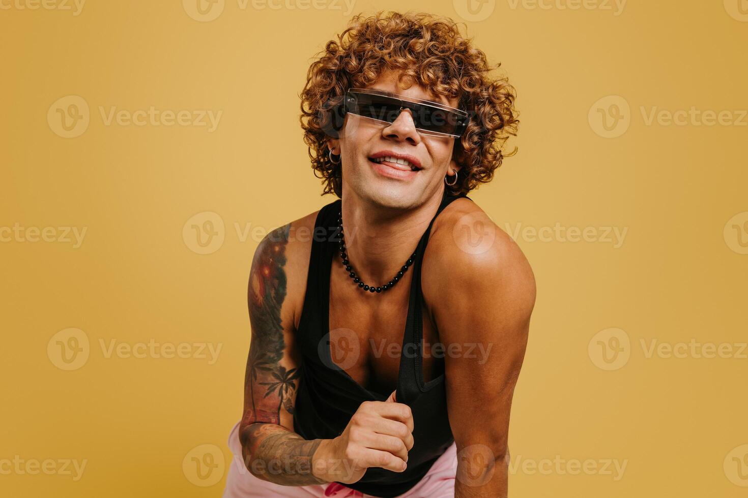 Joyful young gay man in tank top and trendy glasses smiling against yellow background photo