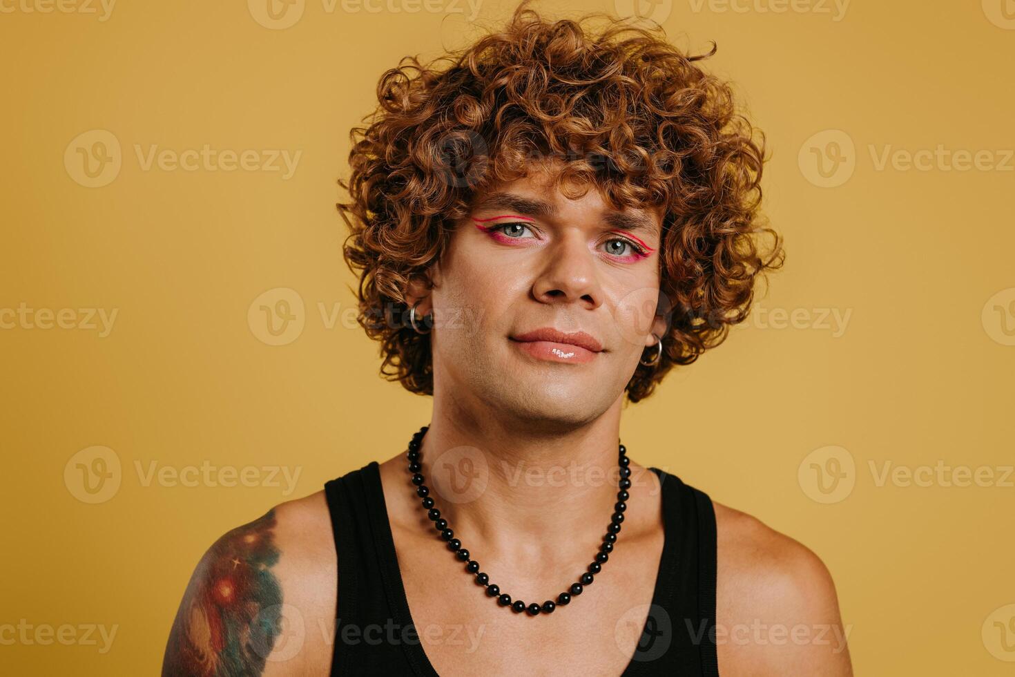 Portrait of handsome young gay man with make-up looking at camera against yellow background photo