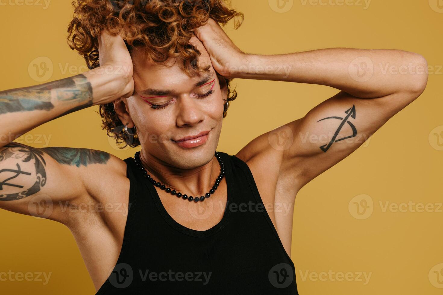 Young curly gay man with make-up holding hands in hair and keeping eyes closed against yellow background photo