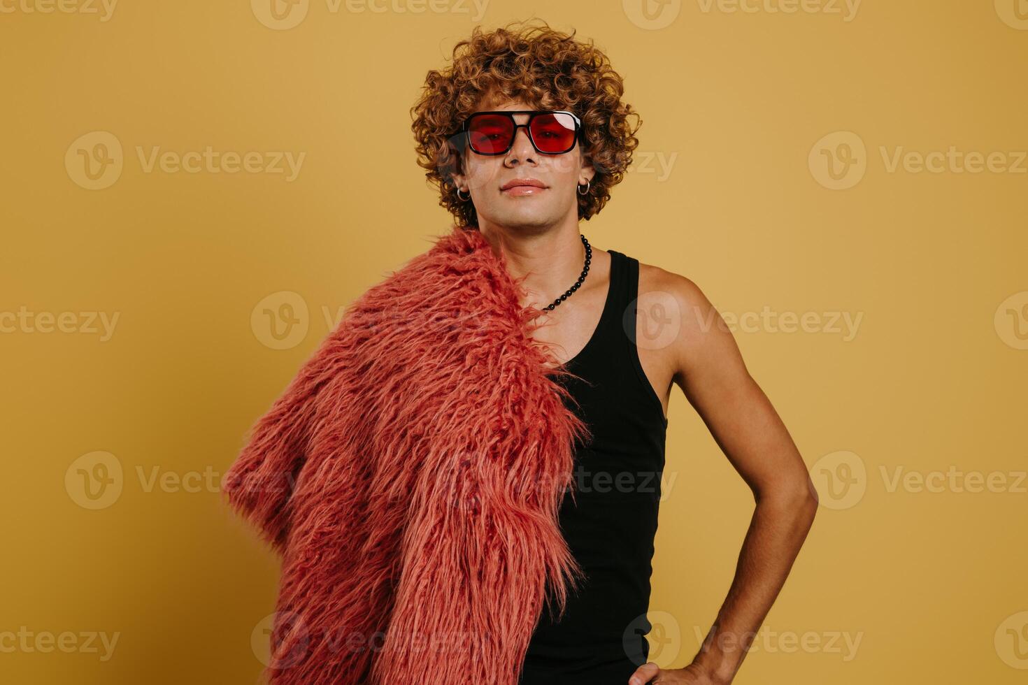 Curly young gay man in trendy glasses carrying fluffy coat on shoulder against yellow background photo