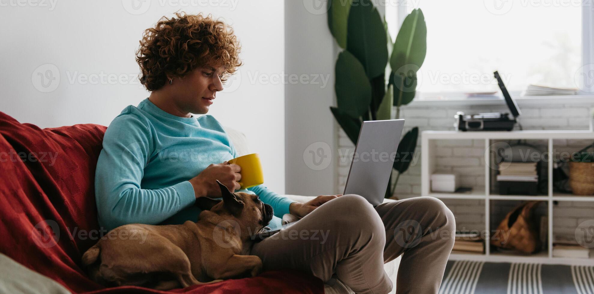 Young man working on laptop and enjoying hot drink while sitting on the couch with his dog together photo