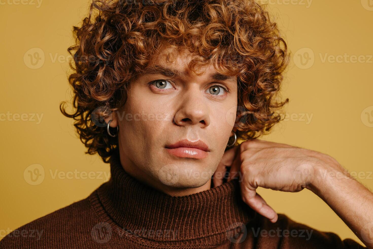 Fashionable curly young man adjusting sweater neck and looking at camera against yellow background photo