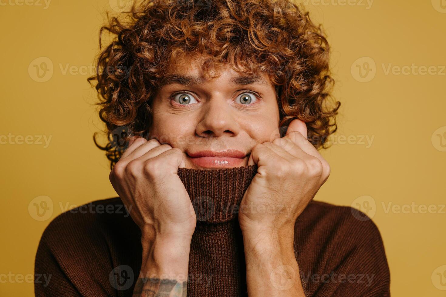 Playful curly young man stretching sweater neck and smiling at camera against yellow background photo