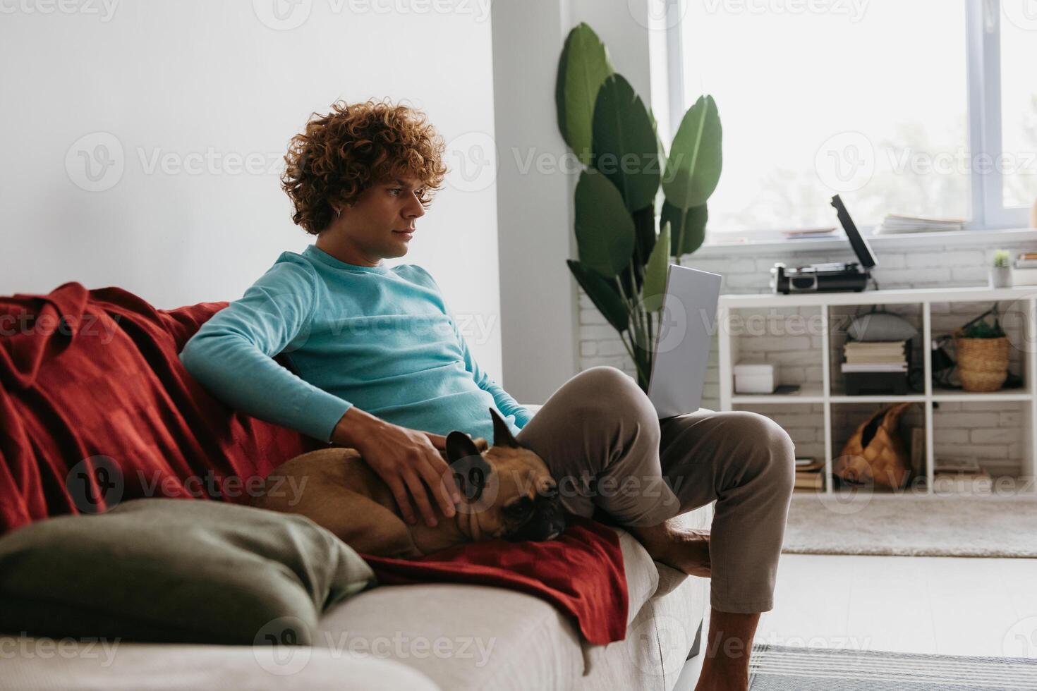 Handsome young curly man working on laptop while relaxing on the couch with his little dog together photo