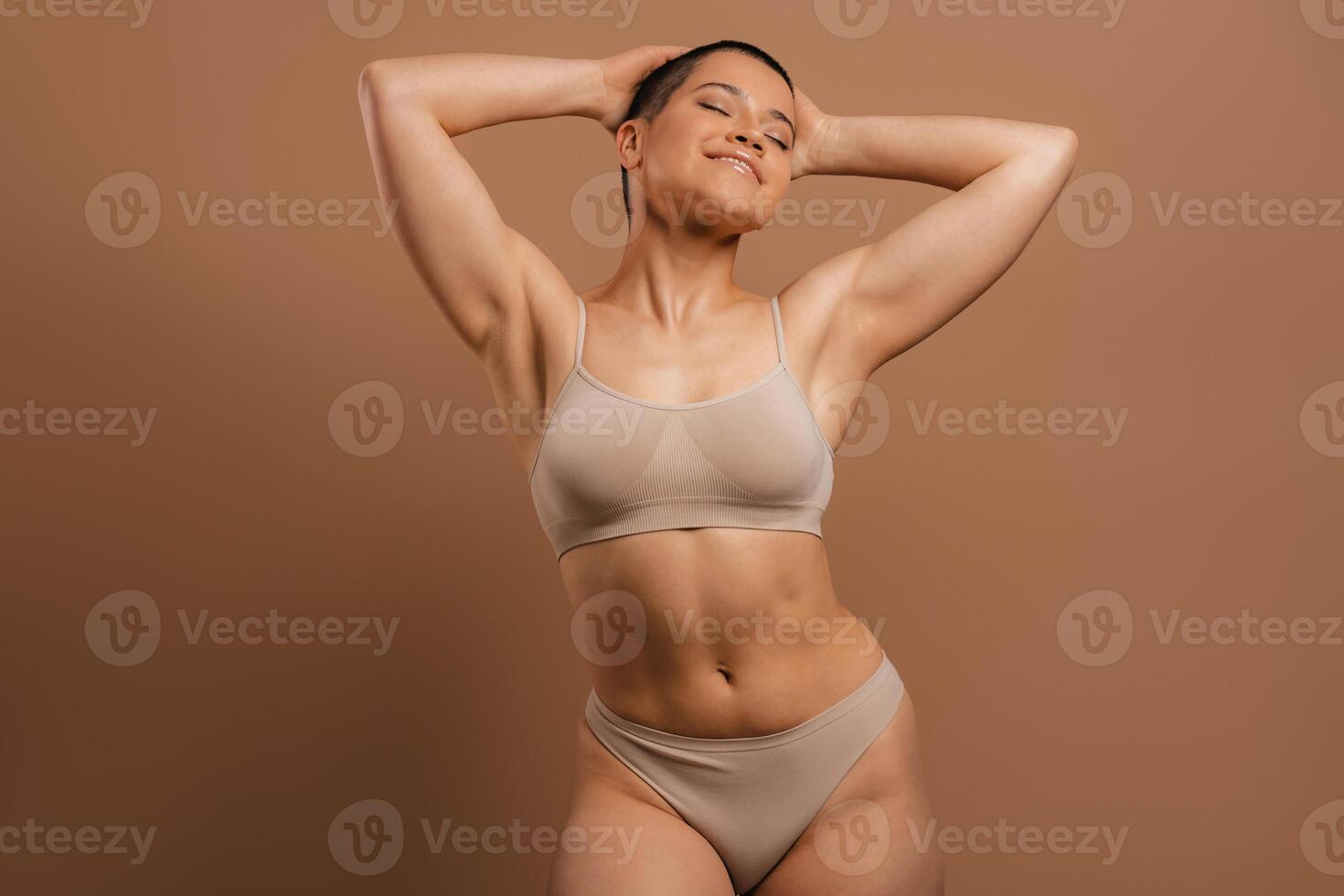 Happy young short hair woman in underwear holding hands behind head against grey background photo
