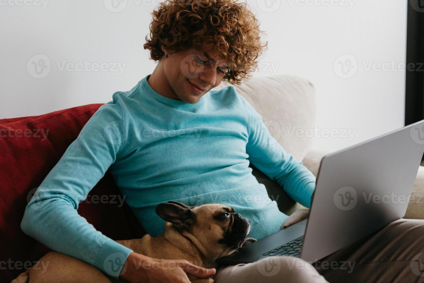 Handsome young curly man petting his dog while working on laptop from home photo