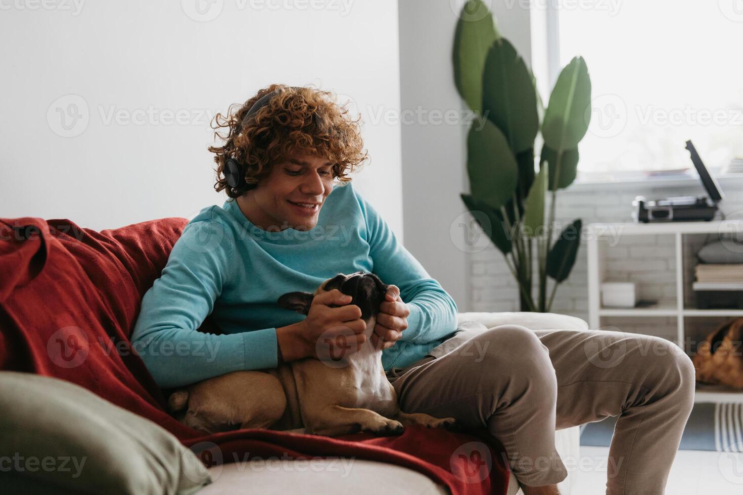 Handsome young curly man in headphones petting cute dog while relaxing on the couch at home photo