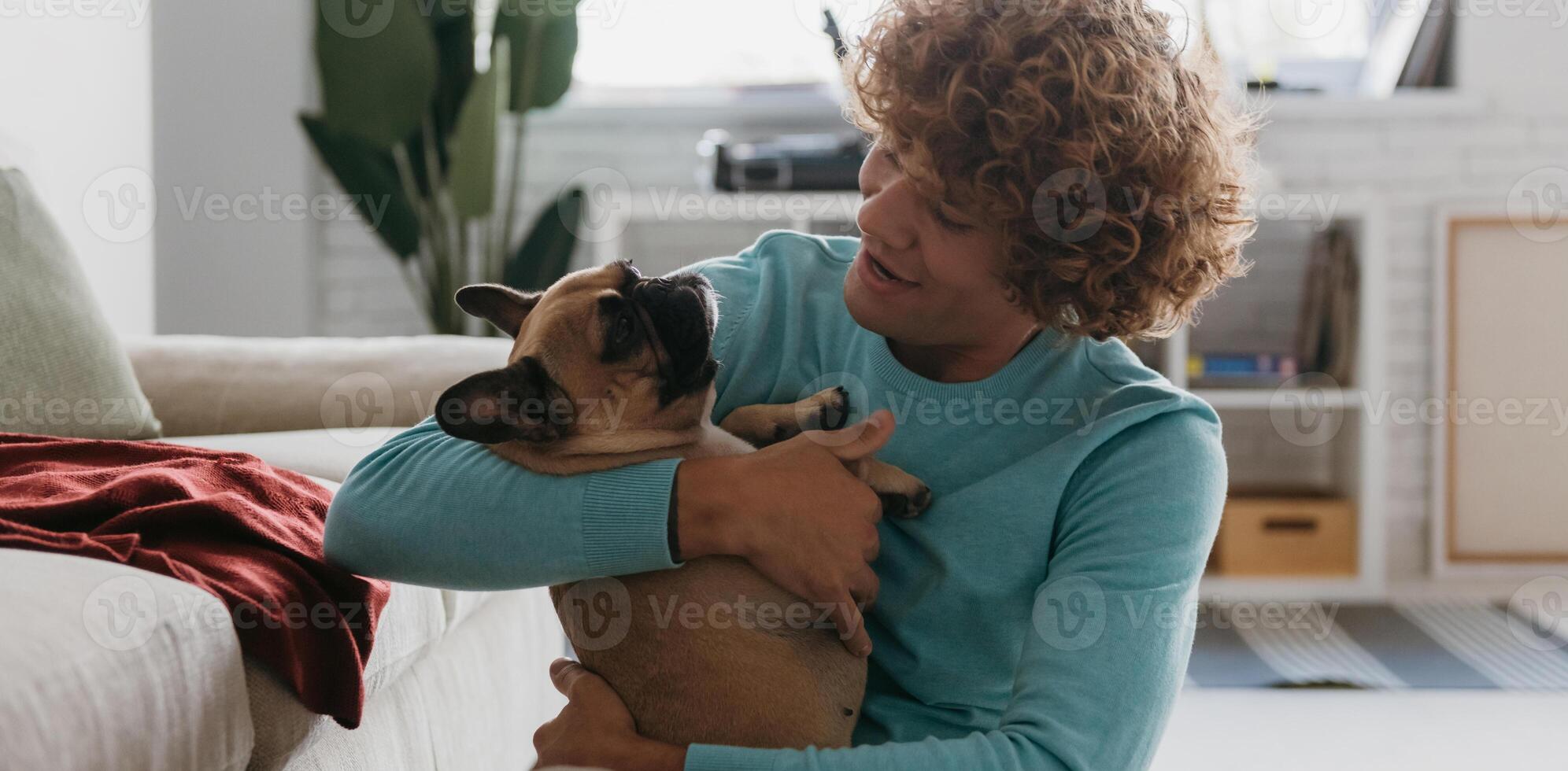 Handsome young curly man holding his cute dog and smiling while relaxing at home together photo